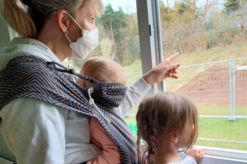 Erzieherin mit Kleinkind im Tragetuch und weiteres Kind neben sich am Fenster der Naturkita Schlüsselau