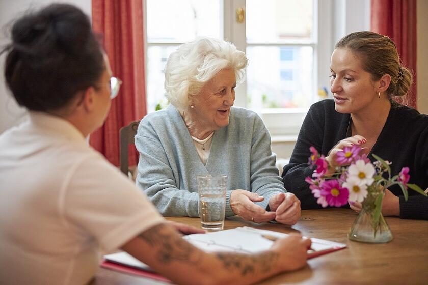 Eine ältere Frau sitzt mit einer Angehörigen und einer Johanniter-Mitarbeiterin an einem Tisch