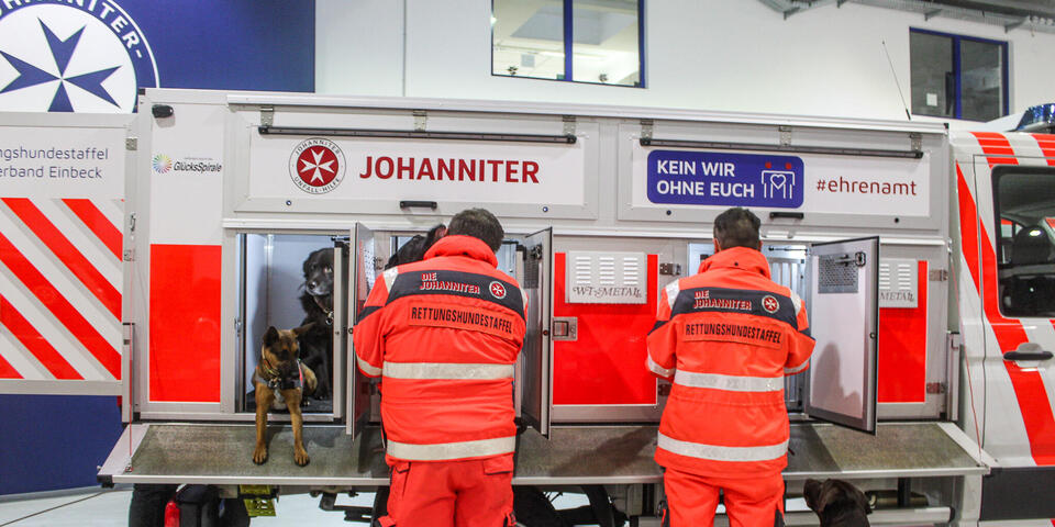 Zwei Johanniter an einem Fahrzeug mit Hundeboxen. Links schauen zwei Hunde aus einer Box