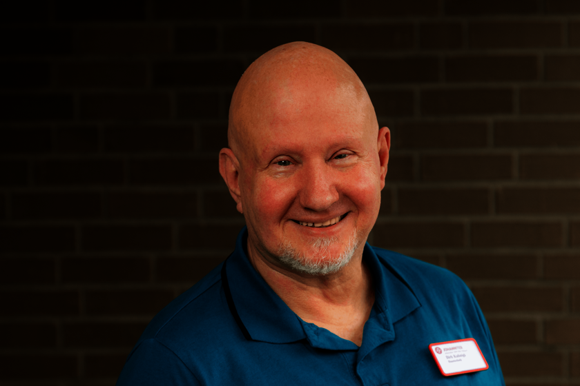 Kopf und Schultern eines kahlköpfigen Mannes mit weißem Kinnbart in einem Poloshirt in Türkis. Er steht vor einer gemauerten Wand und lächelt in die Kamera.