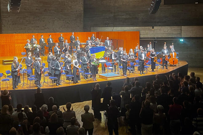 Auf einer Bühne steht ein Orchester, 2 Leute halten eine Ukraine Flagge hoch und das Publikum klatscht.