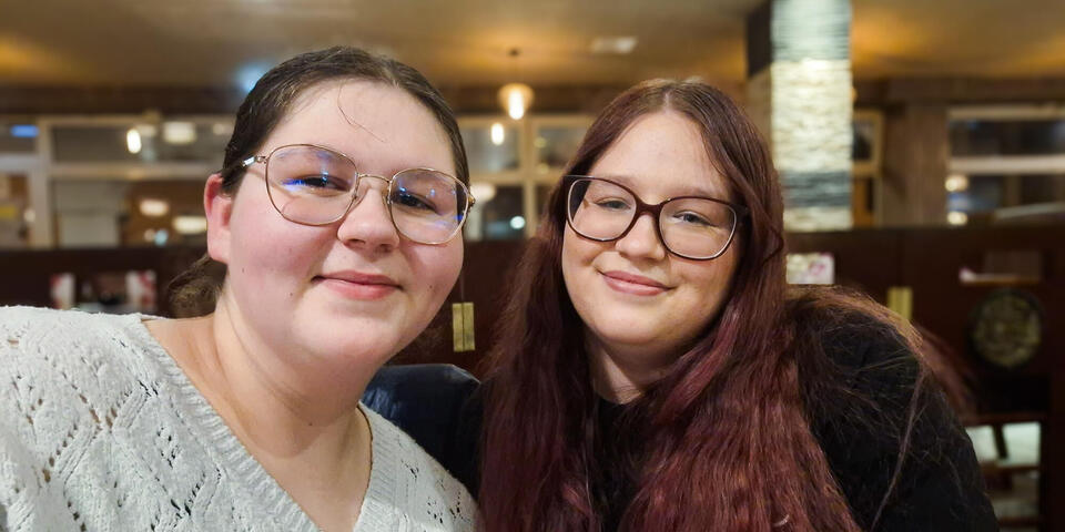Zwei junge Frauen mit braunen Haaren und Brillen lächeln in die Kamera.