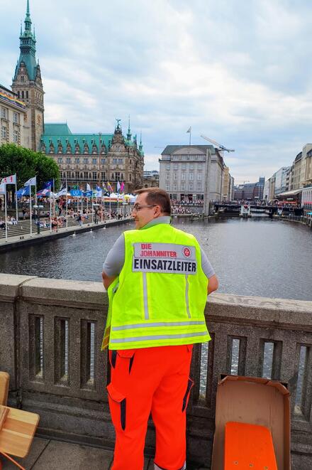 Dirk Willmann steht auf der Brücke an der Binnenalster mit Blick auf das Rathaus. Er trägt eine gelbe Weste mit der Aufschrift Einsatzleiter.