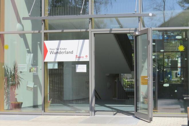 Glaseingang eines Gebäudes mit weit geöffneter Tür und Blick ins Foyer. Auf einem Schild steht „Haus für Kinder, Wunderland“.