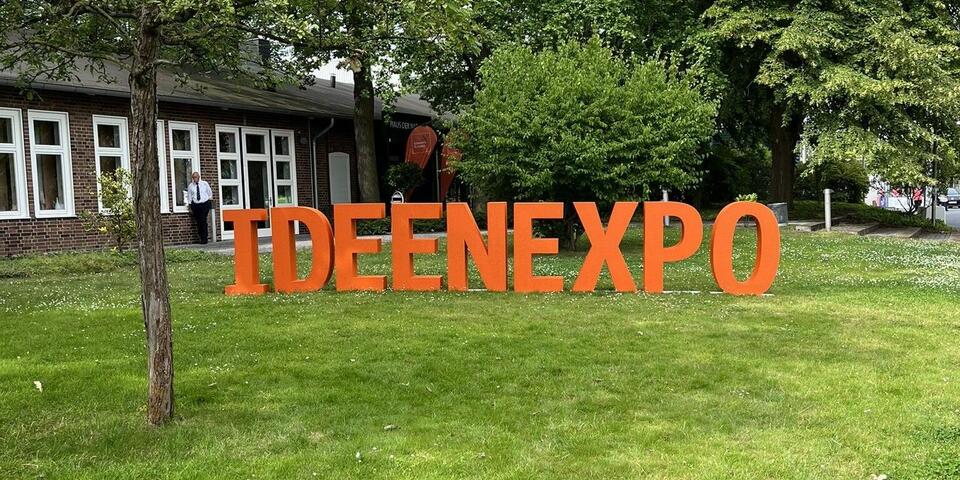 Orangene Großbuchstaben auf einer Wiese. Zu lesen ist "Ideenexpo"