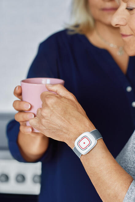 Zu sehen ist eine Hand, die eine Tasse hält. Am Handgelenk das Gerät, um einen Hausnotruf auszulösen