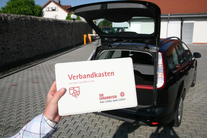 Eine Hand hält einen Johanniter-Verbandkasten vor dem geöffneten Kofferraum eines Autos.