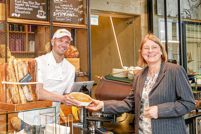 Einer Frau im grauen Blazer wird gerade von einem Bäcker in weißer Arbeitsuniform ein Gebäck überreicht. Sie stehen in einer Bäckerei, er hinter dem Tresen und beide lächeln in eine Richtung.