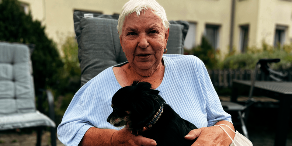Eine Frau mit weißen kurzen Haaren trägt ein blaues Shirt. Auf dem Schoß hält sie einen schwarzen Hund.