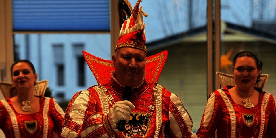 Karnevalsprinz mit  Uniform lächelt in die Kamera, Tänzerinnen im Hintergrund.