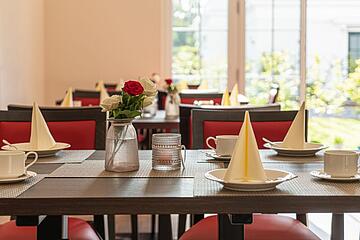 Gedeckter Tisch mit Kaffeegeschirr, Servietten und Blumen in einem hellen Speiseraum.