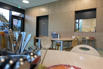 Eine aufgeräumte Cafeteria mit Ausblick auf Küche und Essensausgabe. 