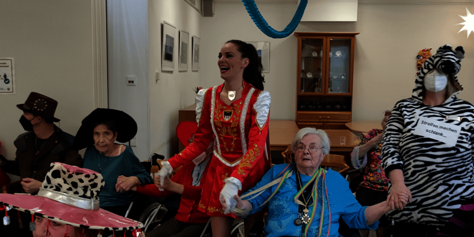 Ein rotgekleidetes Tanzmariechen hält die Hände der neben ihr sitzenden Senioren.