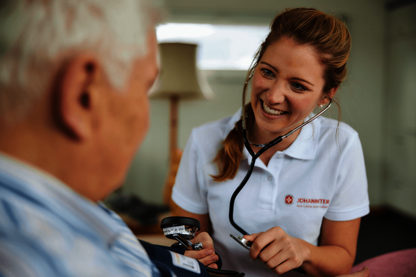 Eine junge Frau in weißem Poloshirt mit Johanniter-Logo spricht lächelnd mit einem älteren Mann und misst ihm den Blutdruck. 
