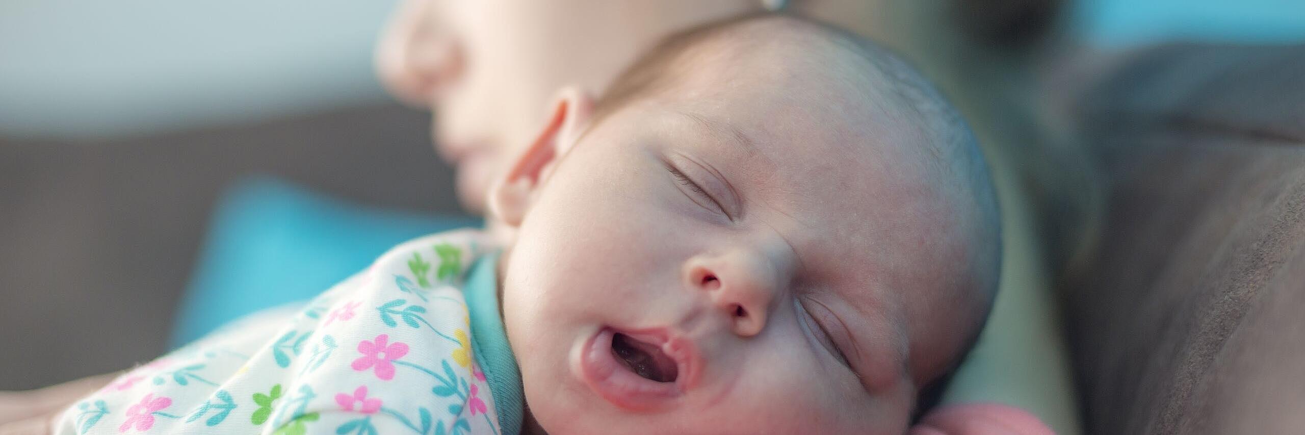 Ein kleines Baby auf dem Arm einer Frau ist auf diesem Bild im Fokus. Das Kind hat die Augen geschlossen und gähnt.