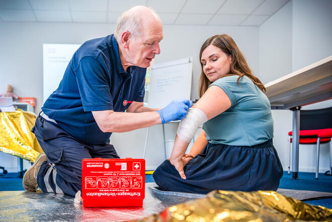 Erste-Hilfe-Trainer mit Handschuhen versorgt Armverletzung einer Teilnehmerin im Kursraum.