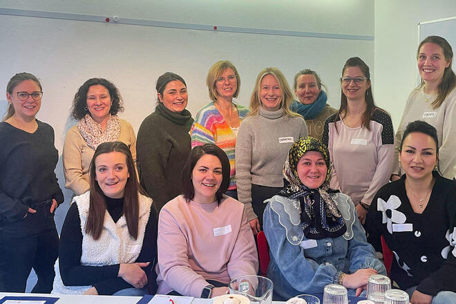 Eine Gruppe von zwölf Frauen steht und sitzt lächelnd in einem Seminarraum vor einer weißen Wand, einige tragen Namensschilder und liegen vor ihnen Notizmaterial und Gläser auf dem Tisch.