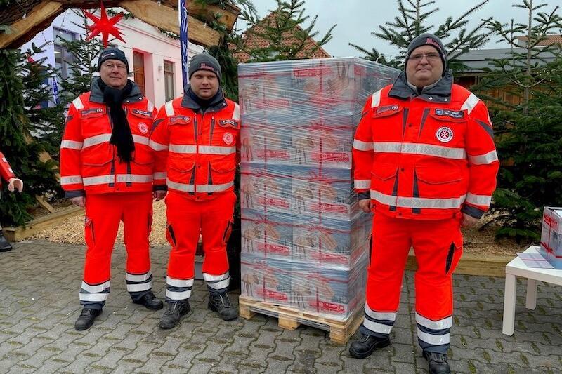 Drei Johanniter Weihnachtstrucker stehen neben einer Palette voller Weihnahtstrucker-Päckchen