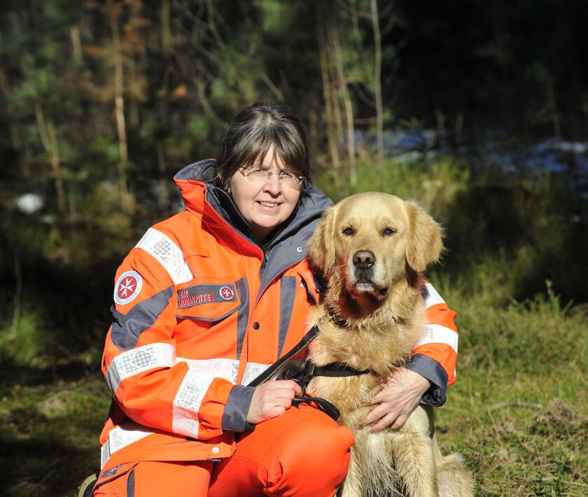 Rettungshundeführerin der Johanniter in Einsatzkleidung kniet auf einer Wiese und lächelt. Neben ihr sitzt ihre hellbrauner Hund, beide sind bereit für den nächsten Einsatz. Im Hintergrund ist ein Wald zu sehen.