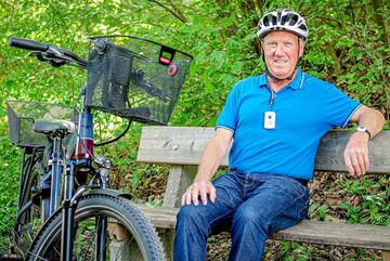 Älterer Mann mit Fahrradhelm sitzt auf einer Parkbank neben einem Fahrrad mit Korb.