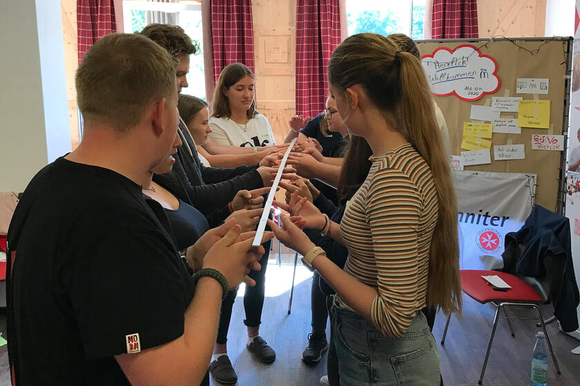 Mehrere Jugendliche stehen in zwei Reihen voreinander und gucken sich an. Sie halten in ihrer Mitte einen Stab, den jede Person mit nur einem Finger trägt.