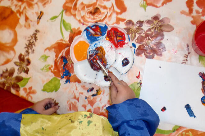 Mit vielen bunten Farben wird ein Ei von der kleinen Hand eines Krippenkindes bemalt.