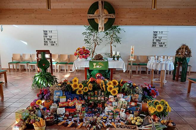 Blick auf einen festlich geschmückten Altarraum einer Kirche. Vor dem Altar ist auf dem Boden eine große Erntedank-Gabenschau aufgebaut: zahlreiche Lebensmittel wie Konserven, Nudeln, Marmeladen und Kekse, umrahmt von Sonnenblumen, bunten Blumensträußen, Kürbissen, Äpfeln, Maiskolben und Körben mit Gemüse.