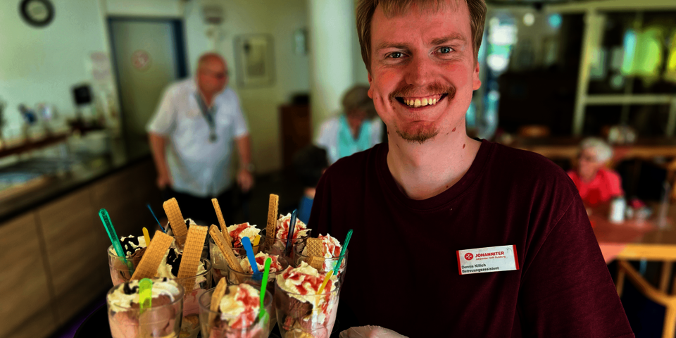 Ein junger Mann in einem dunkelroten T-Shirt mit Johanniter-Namensschild trägt ein Tablett mit Eisbechern