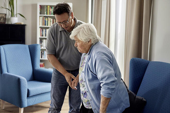 Pflegeszene zu Hause: Ein Mann gibt einer Seniorin Halt beim sicheren Aufstehen.