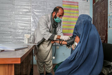 Ein Mann misst den Blutdruck einer Frau, beide tragen eine Mund-Nasen Bedeckung