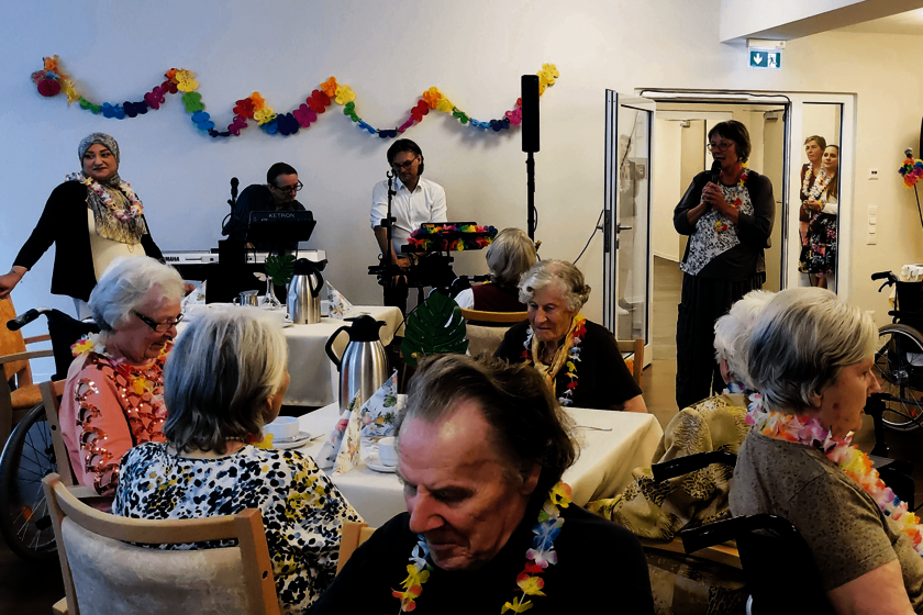 Einige ältere Menschen sitzen an Tischen. Im Hintergrund sind Musiker zu sehen.