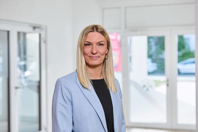 Eine Frau mit hellblonden langen Haaren, einem dunklen Oberteil und einem hellblauem Jackett