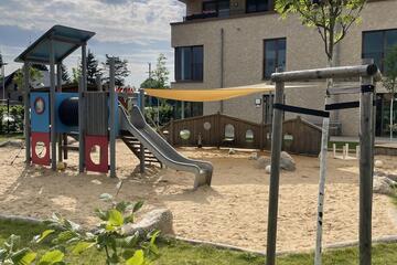Eine Außenansicht des Krippenspielplatzes der Kita Kunterbunt mit Sandkasten, Rutsche und weiteren Spielmöglichkeiten.