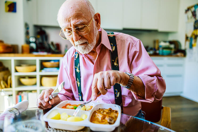 Ein Mann im Seniorenalter schneidet sein Mittagessen aus einer Menübox, während er an einem Esstisch sitzt. Die Mahlzeit besteht aus mehreren Portionen, darunter Gemüse und Kartoffeln.