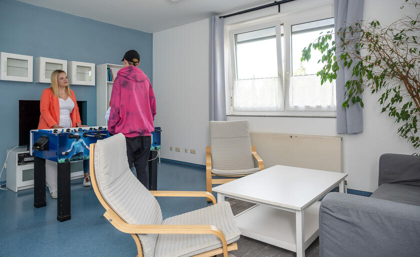 Ein Wohnzimmer mit Kickertisch an dem Menschen spielen. Im Vordergrund zwei beige Sessel, ein weißer Couchtisch und ein graues Sofa. Wände und Boden sind blau.