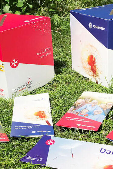 Zwei Spendenboxen der Johanniter mit der Aufschrift Aus Liebe zum Leben, umgeben von mehreren bunten Informationsbroschüren, stehen auf einer Wiese.