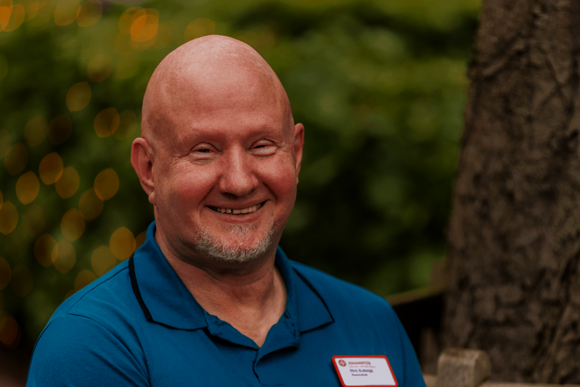 Kopf und Schultern eines kahlköpfigen Mannes mit weißem Kinnbart in einem Poloshirt in Türkis. Er steht im Freien neben einem Baum und lächelt in die Kamera.