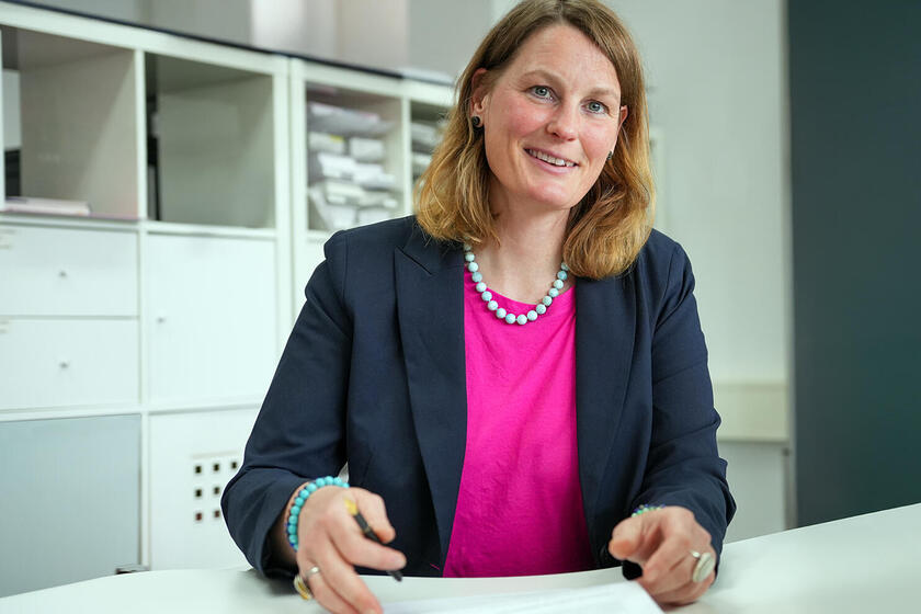 Eine Frau mit blauer Kette und pinkem Oberteil sitzt lächelnd an einem Schreibtisch im Büro und hält einen Stift über ein Dokument, während Regale mit Akten im Hintergrund zu sehen sind.