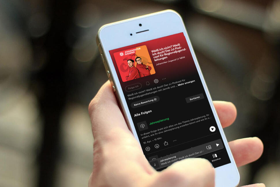 Ein Smartphone in der Hand einer Person. Auf dem Display ist ein Bild von zwei jungen Erwachsenen in Johanniter-Jugend-Begleidung zu sehen, die Rücken an Rücken zueinander stehen. Dies ist das Titelbild des Podcasts.