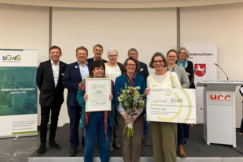 Menschen auf einer Bühne. Im Vordergrund hält eine Frau eine Urkunde hoch, daneben eine Frau mit einem Blumenstrauß und rechts eine Frau mit einem symbolischen Scheck.