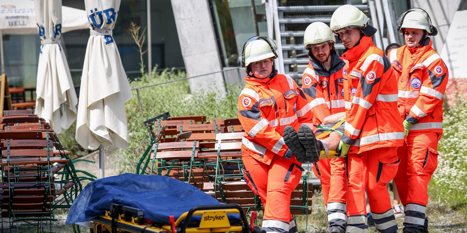 Notfallteam bringt Person auf Trage durch Außenbereich mit Sitzgarnituren.
