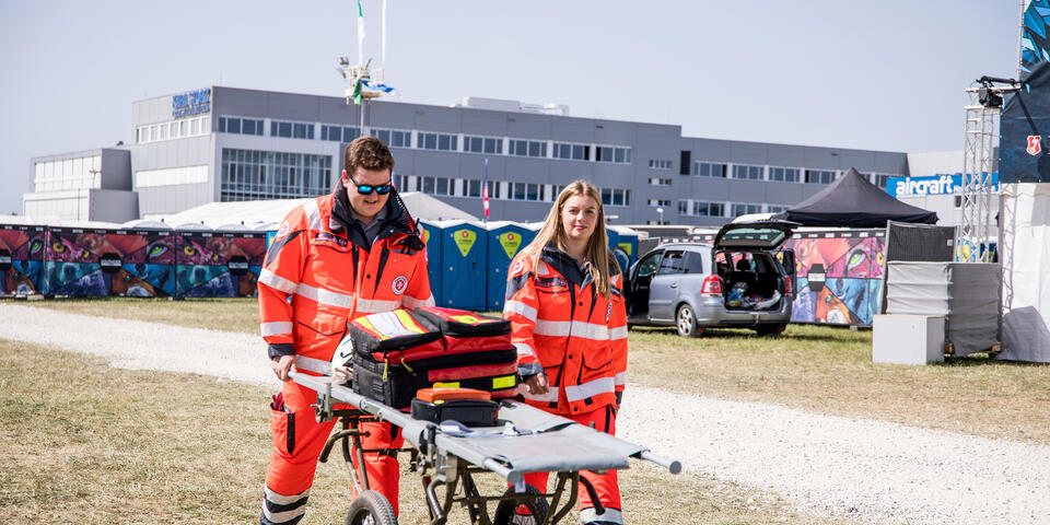 zwei Sanitäter gehen mit Trage und Einsatzrucksack über das Festivalgelände