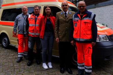 Fünf Ehrenamtliche der Johanniter stehen vor einem Einsatzfahrzeug. Alle tragen verschiedene Johanniter-Einsatz- oder Festkleidung und lächeln.