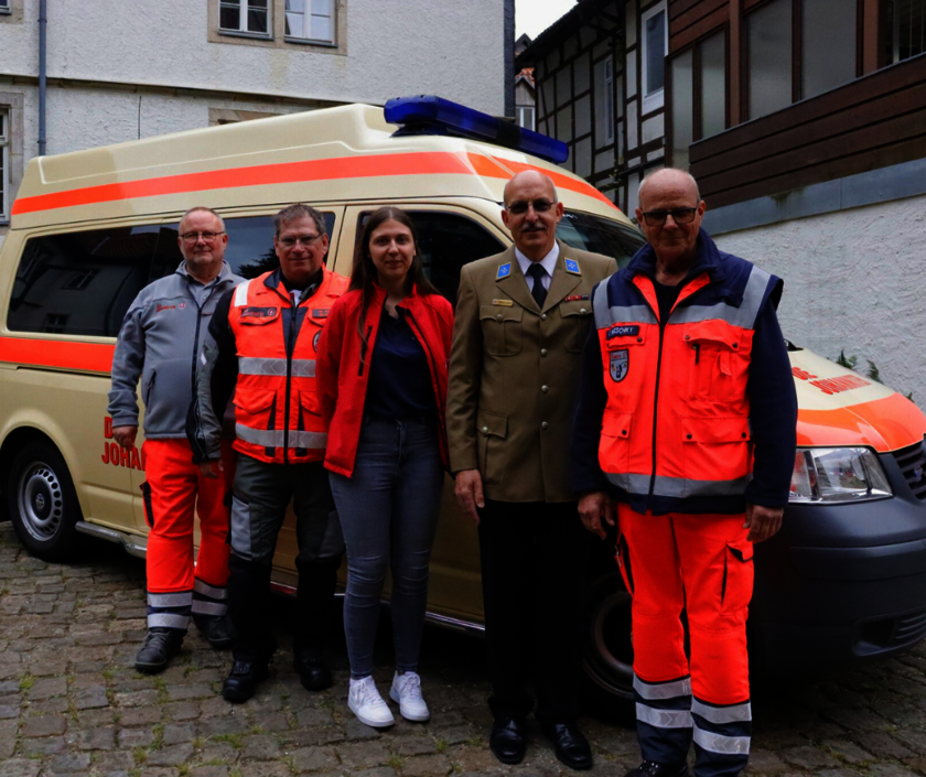Fünf Ehrenamtliche der Johanniter stehen vor einem Einsatzfahrzeug. Alle tragen verschiedene Johanniter-Einsatz- oder Festkleidung und lächeln.