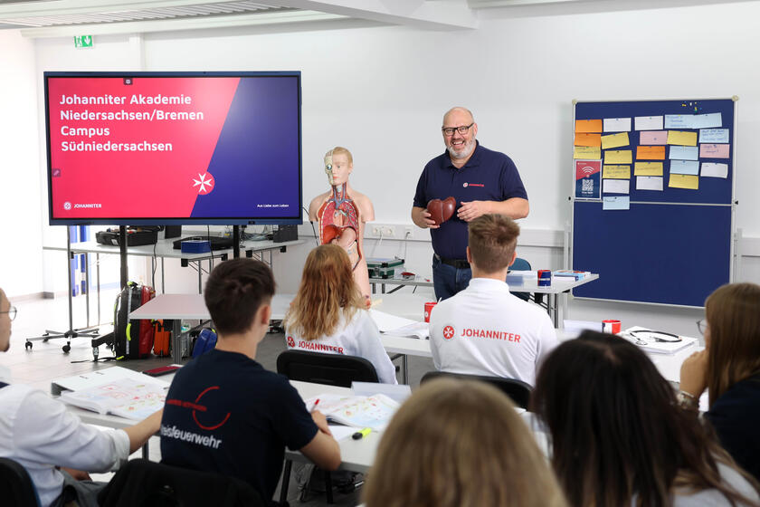 Ein Dozent in Johanniter-Kleidung vor einer Gruppe von Menschen, die an Tischen sitzen.