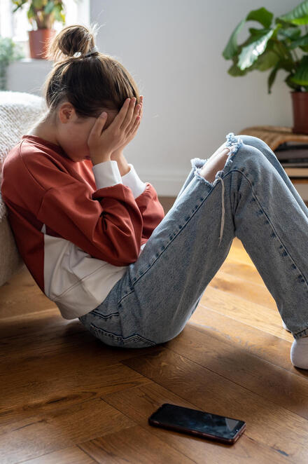 Eine junges Mädchen sitzt auf dem Fußboden, den Rücken ans Bett gelehnt, den Kopf in die Hände gestützt, den Blick nach unten gerichtet. Neben ihr liegt ein Handy auf dem Boden.