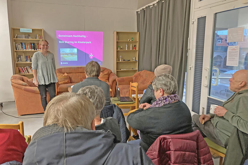 Mehrere Menschen sitzen in Reihen und hören einer Frau bei einem Vortrag zu.