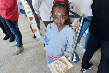 Ein kleines Mädchen mit geschmücktem Gesicht zeigt ihren Teller mit Essen.