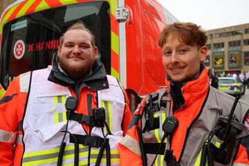 zwei Sanitäter des Johanniter Ortsverbandes Heilbronn, schauen lächelnd in die Kamera beim CSD in Heilbronn. Im Hintergrund steht ein Einsatzwagen der Johanniter. An ihrer Einsatzkleidung sind Funkgeräte befestigt, so sind sie stets abrufbereit.