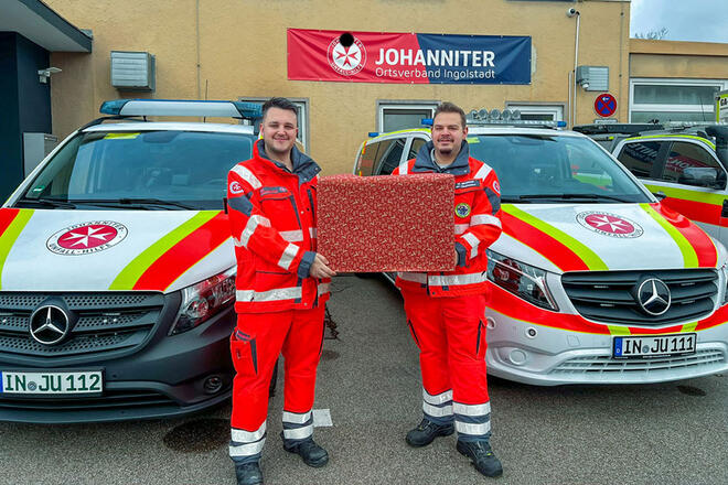 Zwei Johanniter Mitarbeiter stehen zwischen 2 Johanniter Einsatzfahrzeugen und halten ein großes Weihnachtsgeschenk.